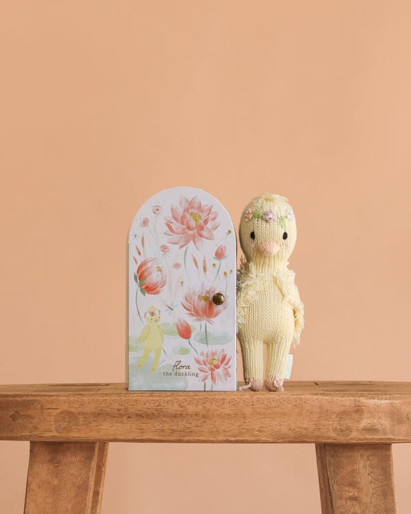 A hand-knit cotton Cuddle + Kind Tiny Flora the Duckling (Mauve) doll, wearing a flower crown, sits on a wooden bench by a floral-decorated door against a peach background.