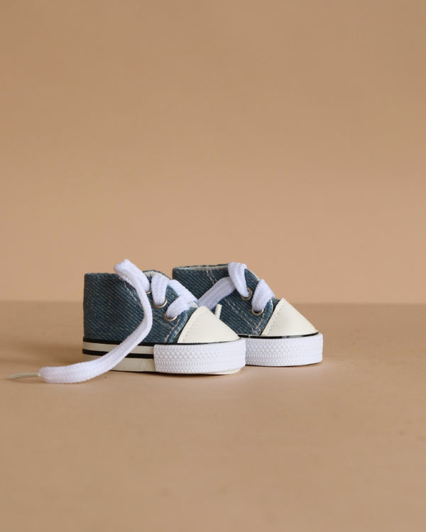 A pair of Minikane Doll Shoes | Converse Style Denim Sneakers with white laces and rubber toes sits on a beige surface with a matching beige background.