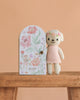 The Cuddle + Kind Tiny Charlotte The Dog, a hand-knit doll in a pink dress, stands beside a floral-patterned door labeled "charlotte the dog" on wood with a peach background. Handmade in Peru, this non-toxic toy is both charming and safe.