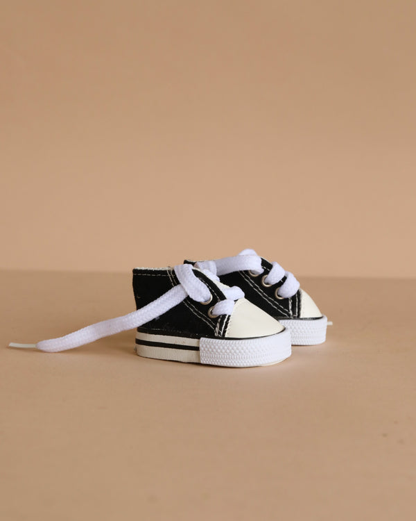 A pair of Minikane Doll Shoes | Converse Style Black Sneakers with white laces are displayed on a beige surface against a beige background.