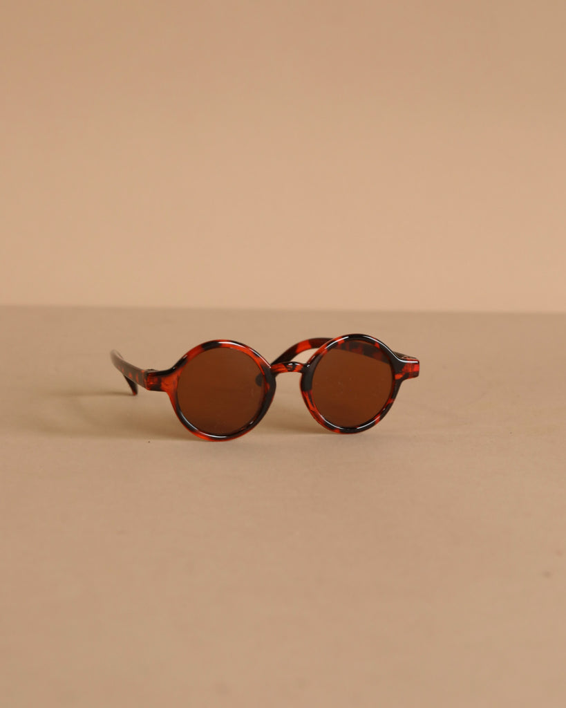 Minikane Doll Accessories | Harry Sunglasses feature round brown lenses and tortoiseshell frames, displayed on a beige surface—perfect stylish accessories for your Minikane doll.