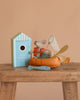 The Maileg Little Brother Gone Fishing Set features a beach mouse with fishing gear, family accessories, and a blue fish beside a blue-and-white striped beach house toy, all displayed on a wooden table against a beige background.