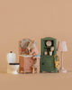 The Maileg Little Sister Bathroom Set features two Little Sister Mouse figures at a dollhouse sink with mirror, plus a toy toilet, green cabinet with clock, purse, toiletries, and a patterned lamp—all set against a plain light brown background.