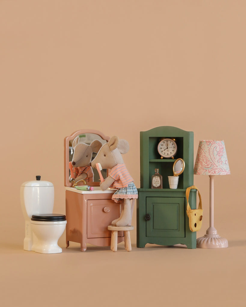 The Maileg Little Sister Bathroom Set features two Little Sister Mouse figures at a dollhouse sink with mirror, plus a toy toilet, green cabinet with clock, purse, toiletries, and a patterned lamp—all set against a plain light brown background.