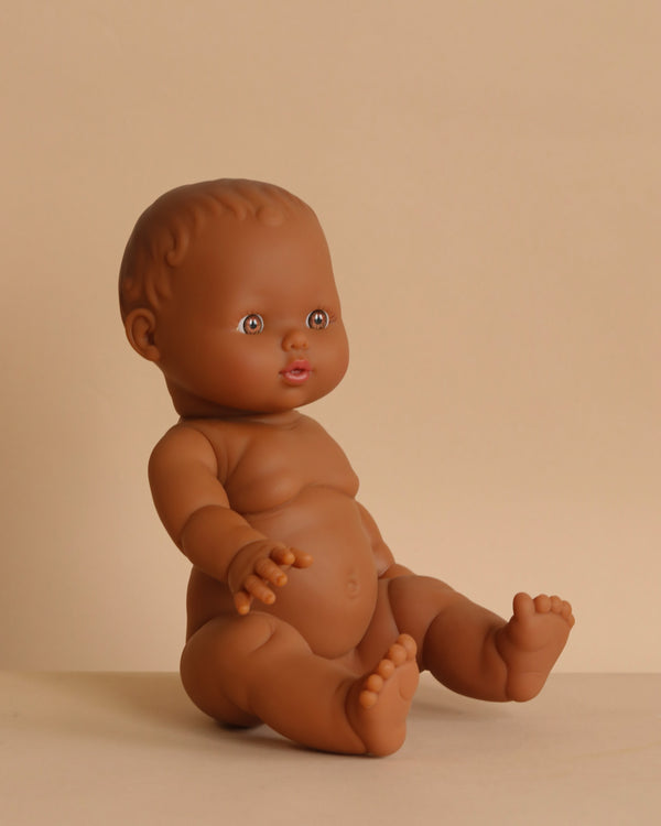The Minikane Doll (13") - Leonie features brown skin and short hair, sits upright on a light beige surface with extended arms and legs, and is made from phthalate-free vinyl for safe play. Clothing sold separately.