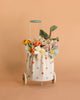 The Basket of Felt Flowers features a small Maileg trolley basket with a white polka-dot bag, filled with a colorful wool felt bouquet, set against a plain peach background.