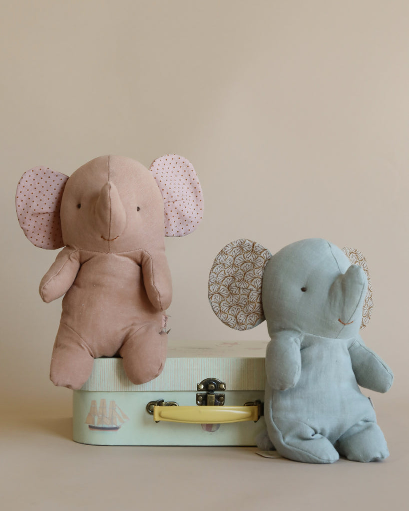 Two Maileg Small Elephant toys, one pink and one gray, crafted from the softest fabric, sitting upright on a small suitcase against a plain beige background.