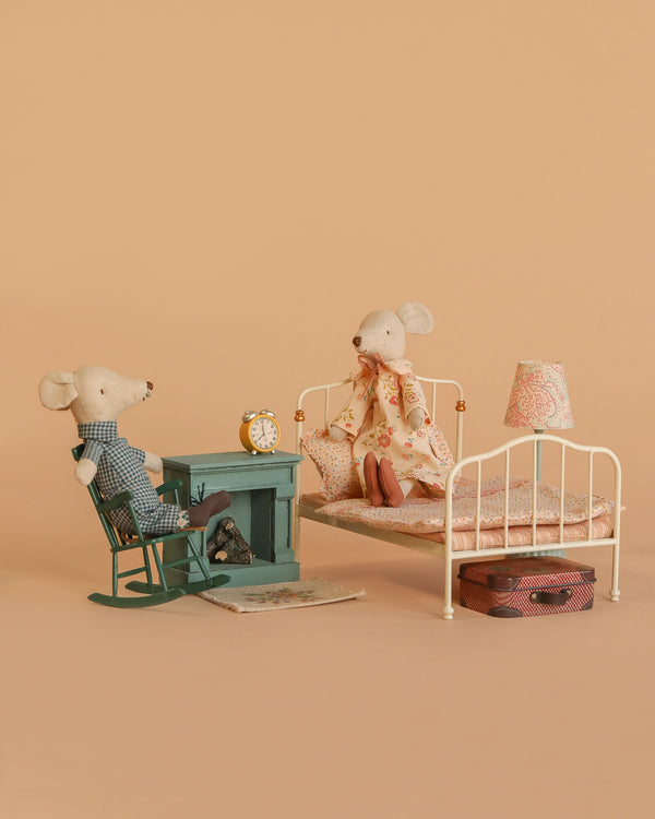 The Maileg Grandma and Grandpa Bedroom Set features two mice relaxing in a cozy miniature room with vintage furniture and charming accessories like a rocking chair, fireplace, bed, lamp, clock, rug, and suitcase against a beige background.
