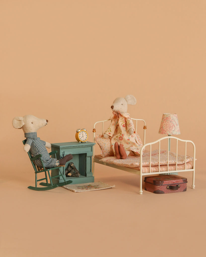 The Maileg Grandma and Grandpa Bedroom Set features two mice relaxing in a cozy miniature room with vintage furniture and charming accessories like a rocking chair, fireplace, bed, lamp, clock, rug, and suitcase against a beige background.