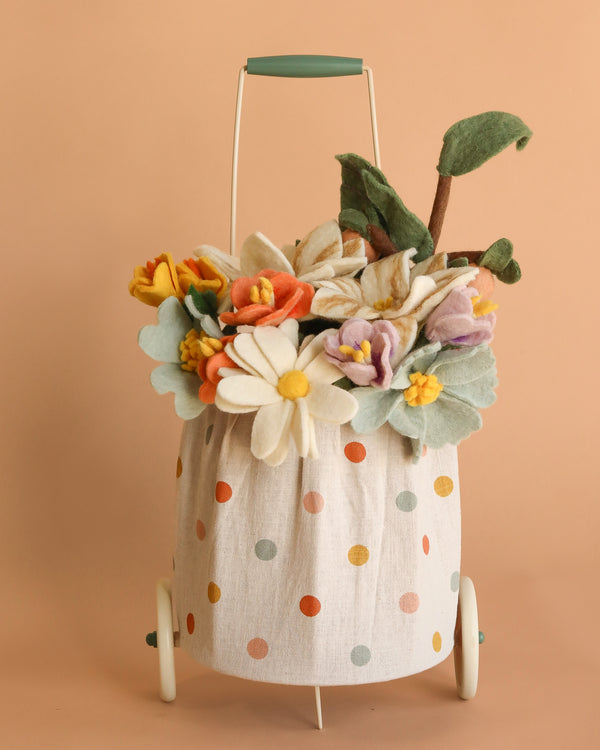 The Basket of Felt Flowers features a cheerful wool felt bouquet with colorful flowers and green leaves in a Maileg trolley basket, all set against a soft peach background.