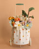 The Basket of Felt Flowers features a cheerful wool felt bouquet with colorful flowers and green leaves in a Maileg trolley basket, all set against a soft peach background.