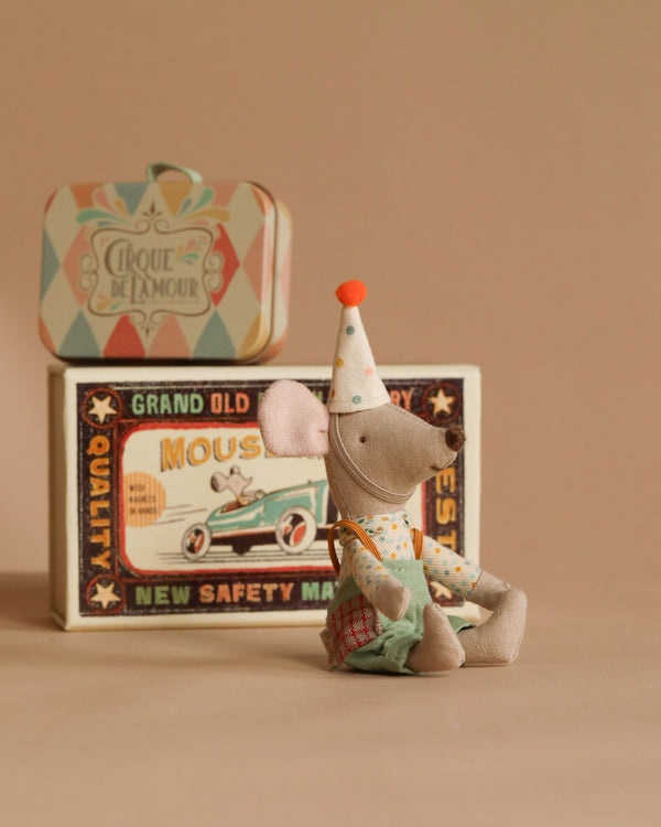 A small fabric mouse, Maileg Little Brother Mouse, wears charming clown clothes and a polka dot outfit. It sits in front of a decorated vintage circus-themed box featuring a car and the words "Cirque de l'amour," "Grand Old Quality," and "Mouses," enchanting all who see it.