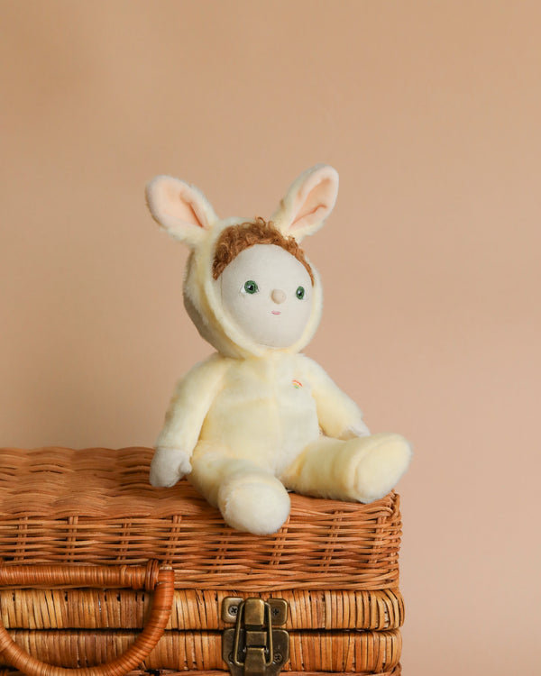 A Dinky Dinkums - Bunny plush toy sits on top of an Easter basket against a soft peach background. The toy is pale yellow with small, green eyes and curly hair.