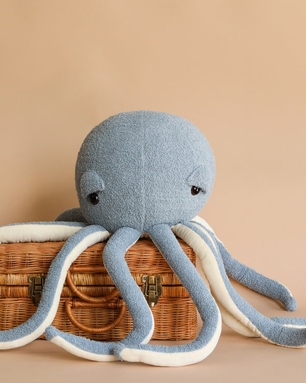 A Octopus Stuffed Animal sits on a wicker basket against a soft beige background, with its tentacles draped over the basket's edges, perfect as a children's room decoration.
