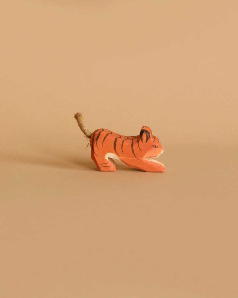 A small handcrafted wooden figurine of an orange tiger with black stripes and a rope tail, named Ostheimer Tiger Lurking, Small, is placed against a beige background. The toy appears to be in a crouching or playful position, perfect for imaginative play.