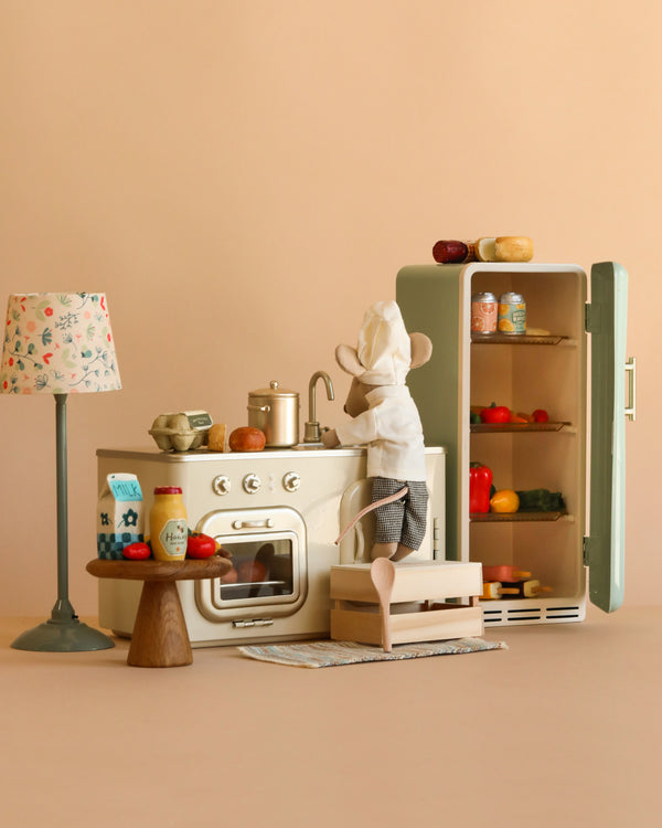 A Chef Mouse stands at the Maileg Little Chef’s Kitchen with a stove, sink, and fridge. Toy food, kitchenware, a lamp, and a small table complete this adorable cooking scene.