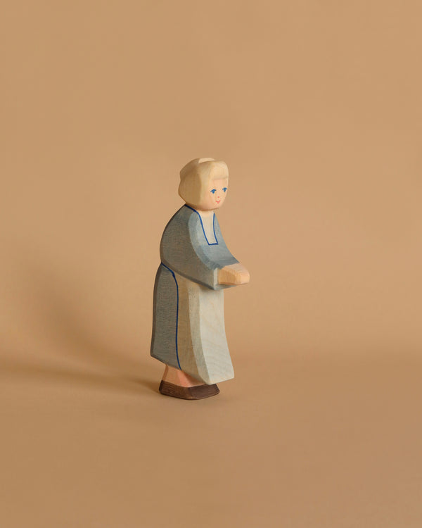 The Ostheimer Grandmother, a handcrafted wooden figure with light hair, dressed in a blue robe and white apron, stands on a beige background. The figure features simplified facial characteristics and appears to be leaning slightly forward, making it perfect for imaginative play.