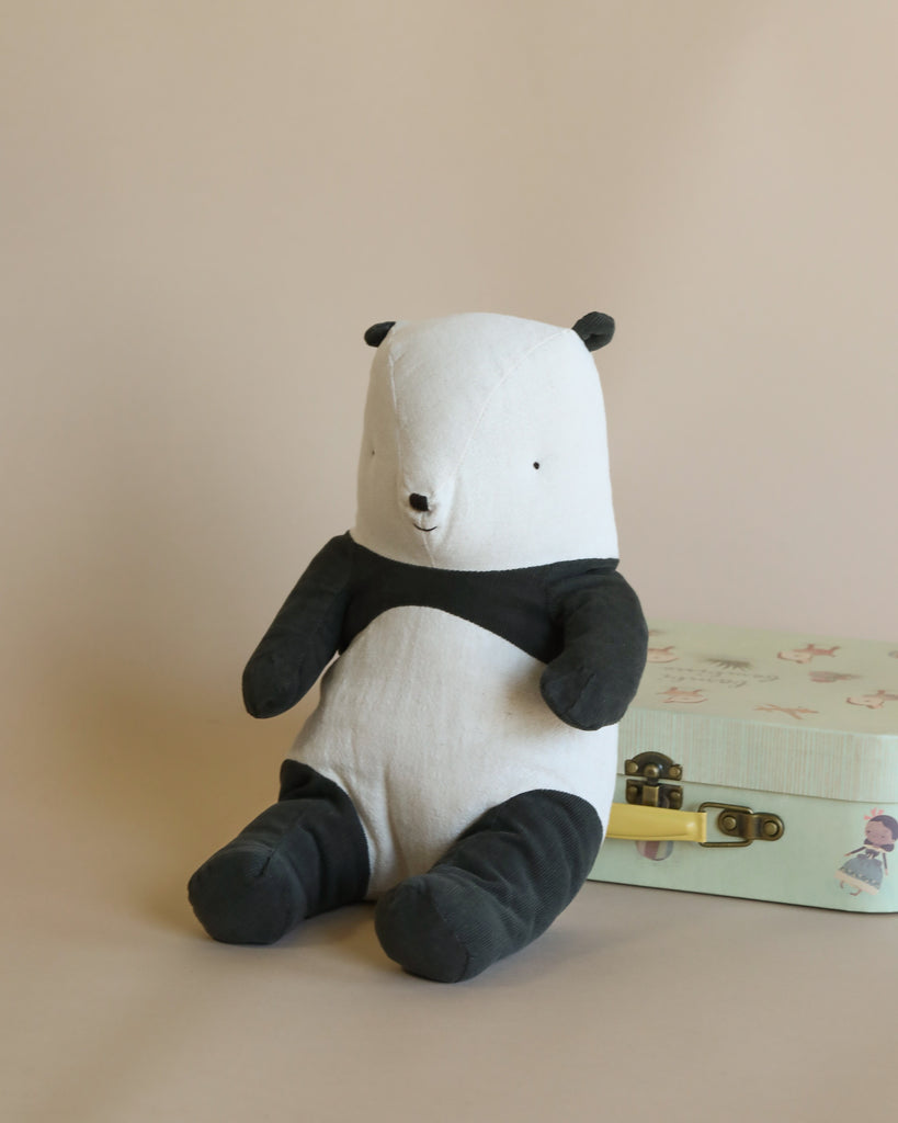 A Maileg Panda Stuffed Animal sits upright against a pastel pink background, accompanied by a closed pastel suitcase with floral patterns. The panda has a simple, charming design with black and white colors.