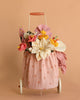The Basket of Felt Flowers, a small wheeled cart with a pink polka-dot fabric bag filled with colorful wool felt flowers, stands against a plain peach background.