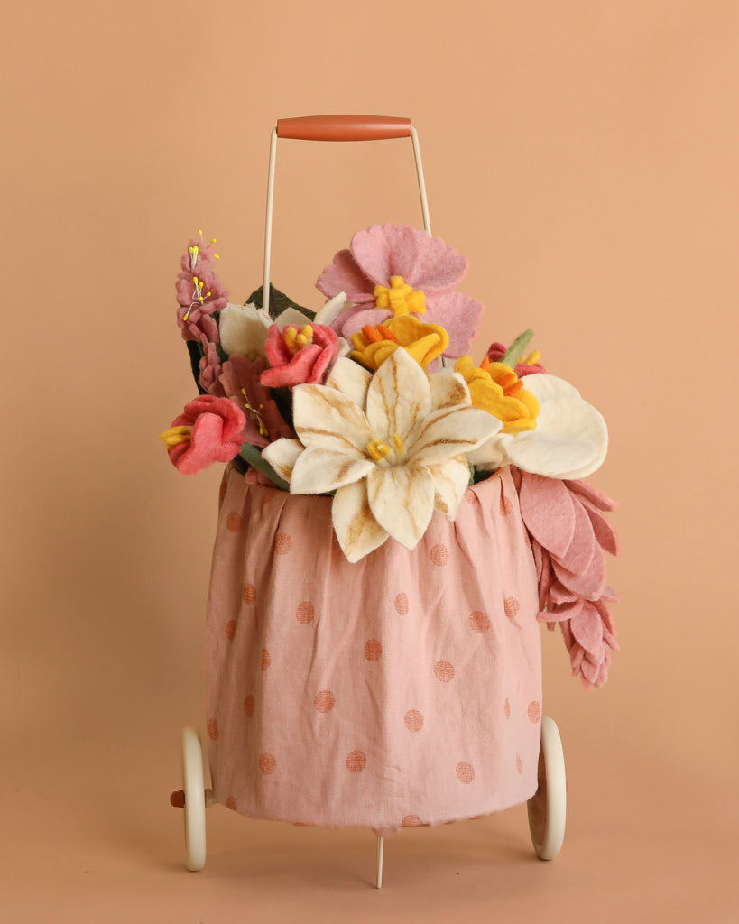 The Basket of Felt Flowers, a small wheeled cart with a pink polka-dot fabric bag filled with colorful wool felt flowers, stands against a plain peach background.