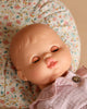 The Minikane Doll with Sleeping Eyes (13") - Lily With Clothing rests on a floral cushion, her eyes closed and pink lips visible, dressed in a light pink outfit with a button.