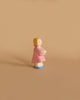 The Ostheimer Daughter in Pink Dress is a small wooden figure of a girl with short blonde hair, wearing a pink dress and blue shoes, holding a white object in front of her. Set against a plain, light brown background, this handcrafted toy from the Ostheimer brand encourages imaginative play in children.