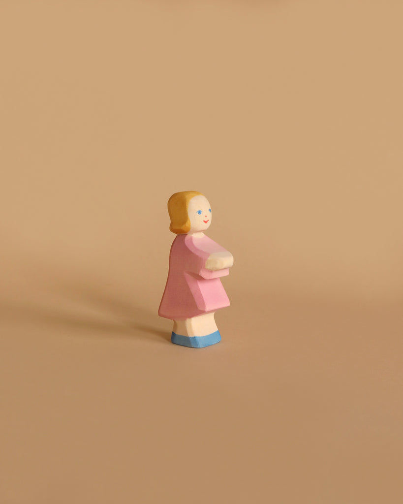 The Ostheimer Daughter in Pink Dress is a small wooden figure of a girl with short blonde hair, wearing a pink dress and blue shoes, holding a white object in front of her. Set against a plain, light brown background, this handcrafted toy from the Ostheimer brand encourages imaginative play in children.