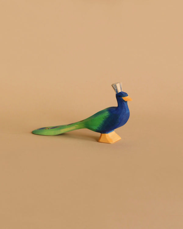 The Ostheimer Peacock, a colorful wooden bird figurine handcrafted from sustainably sourced materials, features a blue body, green tail, small yellow beak, and a decorative crest on its head. It stands against a plain beige background.