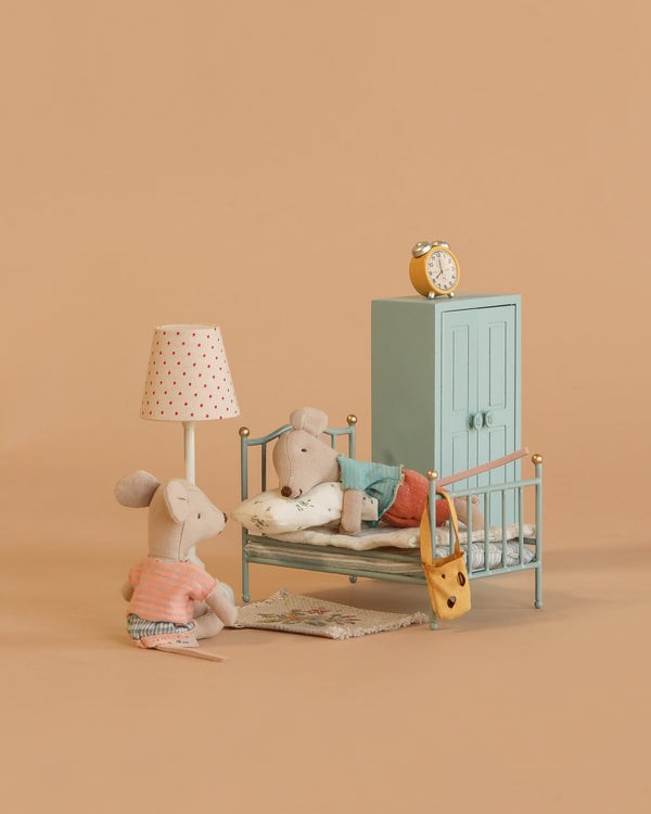 Two Maileg mice from the Little Brother and Sister Bedroom Set create a cozy scene: one sits by a vintage bed as the other is tucked in. The Maileg room also features a lamp, rug, wardrobe, and clock for added charm.