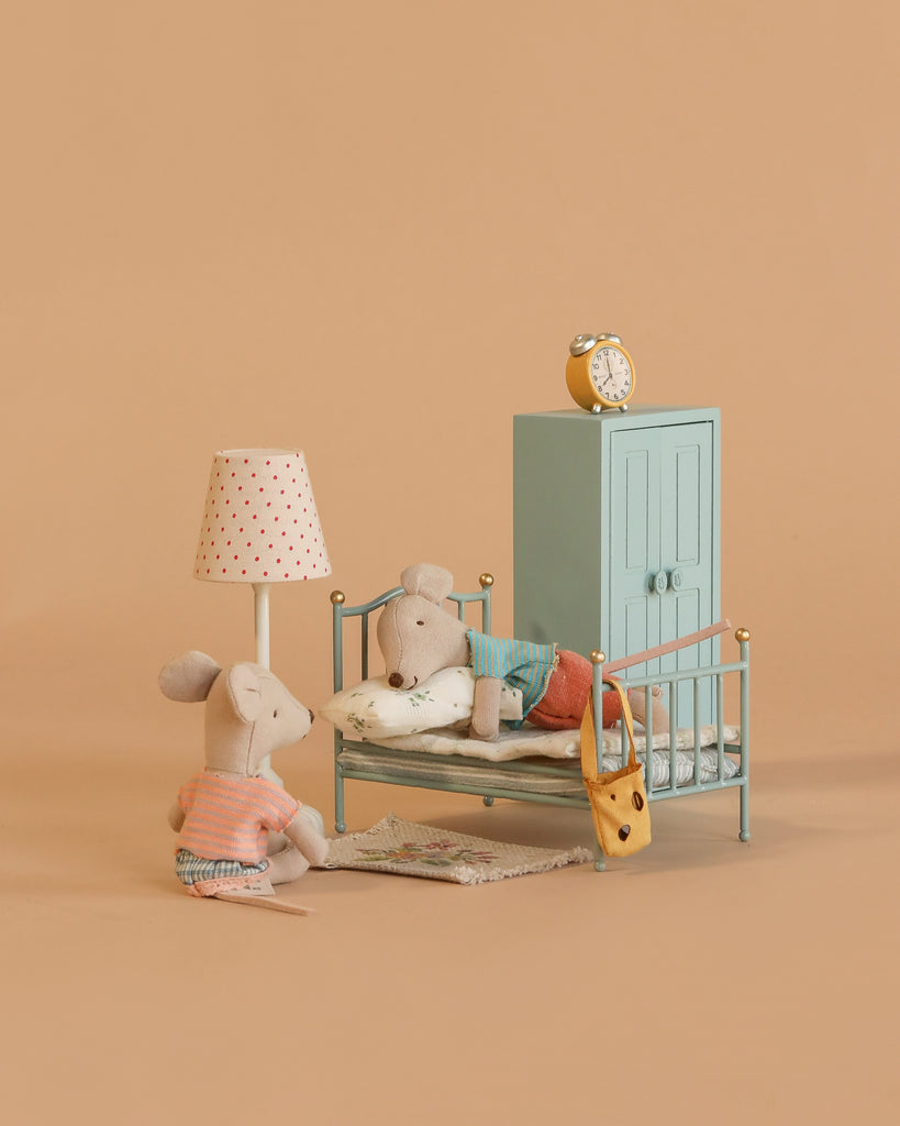 Two Maileg mice from the Little Brother and Sister Bedroom Set create a cozy scene: one sits by a vintage bed as the other is tucked in. The Maileg room also features a lamp, rug, wardrobe, and clock for added charm.