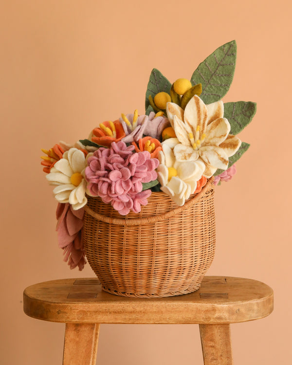 The Basket of Felt Flowers, filled with eco-friendly felt blooms and vibrant leaves, rests on a wooden stool against a peach-colored background.