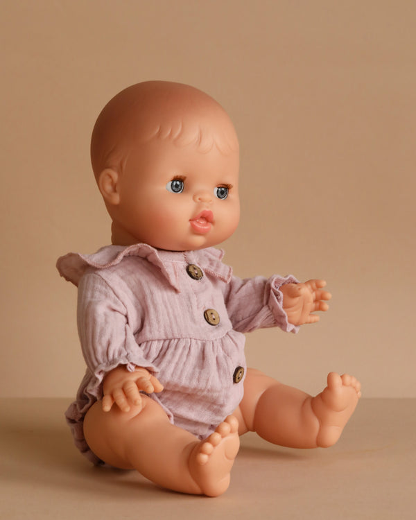 The 13" Minikane Doll with Sleeping Eyes – Lily, dressed in a pale pink romper with large brown buttons, sits upright on a beige surface against a beige background.