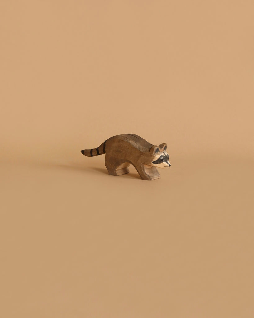 The Ostheimer Raccoon Running, a small hand-carved wooden figurine, is perfect for imaginative play. This raccoon features distinct facial markings and a bushy, striped tail. It stands on a plain brown surface against a solid light brown background, creating a minimalist and earthy aesthetic reminiscent of Ostheimer toys.