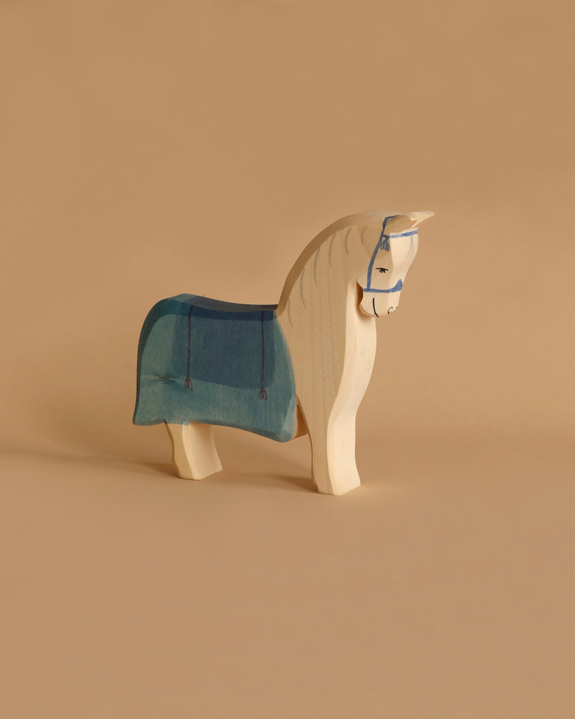 An Ostheimer Horse with Saddle, showcasing a simple and smooth design, stands on a tan background. This light-colored wooden toy features a painted blue harness on its head and a blue-and-green cape-like covering on its back. Handcrafted from sustainably sourced materials, this wooden horse exemplifies the quality of Ostheimer toys.