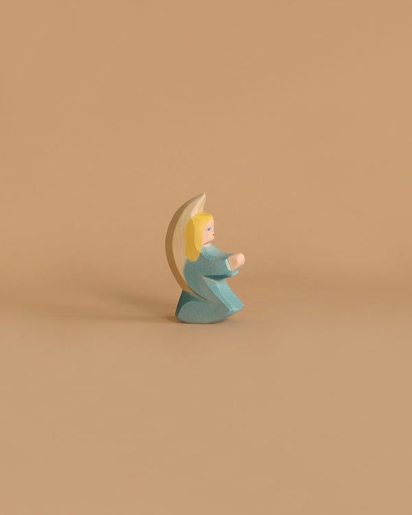 Ostheimer Little Angel - Turquoise: This wooden figurine features an angel with blond hair, donned in a turquoise robe and white wings, kneeling with hands clasped. Handcrafted from sustainably sourced materials, the piece embodies the timeless quality characteristic of Ostheimer wooden toys. The background is plain white.