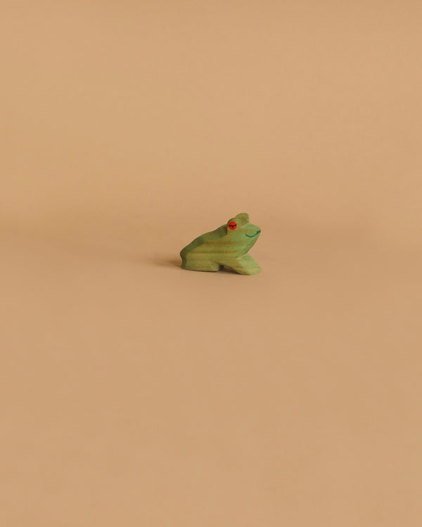 A small Ostheimer Frog Sitting figurine, painted green with red eyes, is centrally placed on a plain, light brown background. The handcrafted design of the Ostheimer toy is simple and minimalistic, adding a vibrant touch of color to the neutral setting.