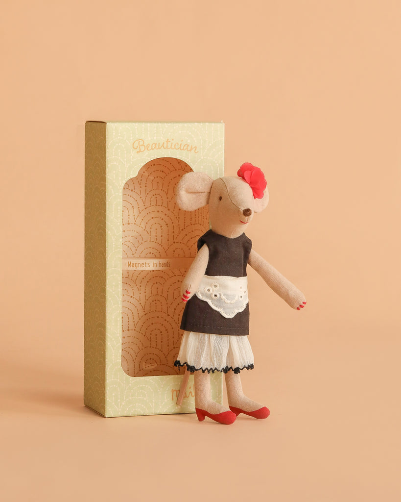 The Maileg Beautician Mouse, Mum wears a black dress, white apron, red shoes, and a red flower headpiece. She stands beside a beige "Beautician" box, ready to inspire endless imaginative dress-up play.
