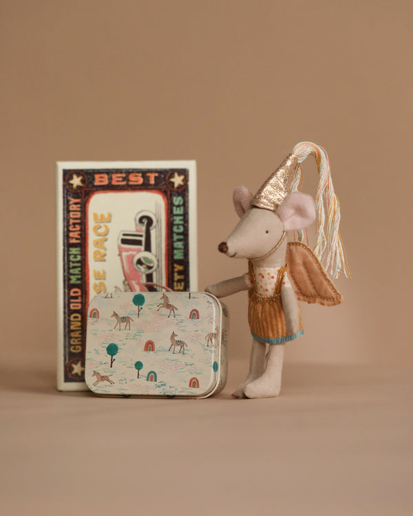 The Maileg Little Sister Mouse toy stands in a whimsical fairy outfit with a party hat and golden wings beside a matchbox bed, holding an illustrated tin suitcase featuring delightful animal designs, set against a plain brown background.