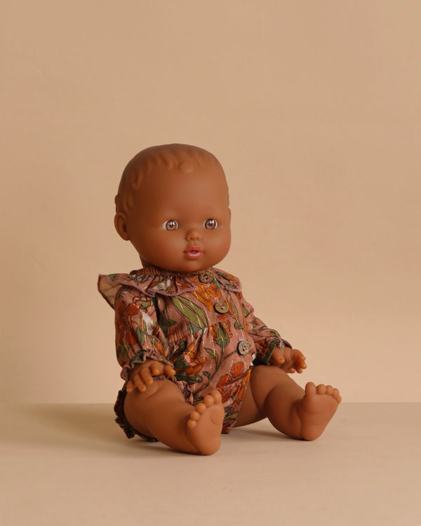 The Minikane Doll (13") - Leonie With Clothing features a brown-skinned baby doll with short hair sitting on a beige surface, dressed in a patterned brown and green romper with ruffled collar and buttons against a plain beige background.