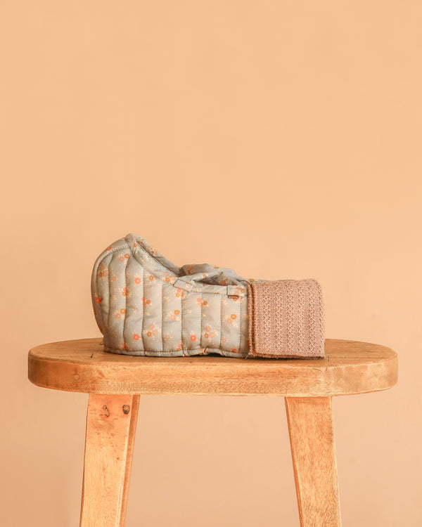 The Maileg Carry Cot, Large - Blue, featuring a quilted floral pattern and knitted strap, sits atop a wooden stool against a plain beige backdrop.