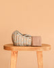The Maileg Teddy Bear in Carry Cot, featuring a soft floral-patterned carrier with a knitted panel, is displayed on a wooden stool against a simple beige background.