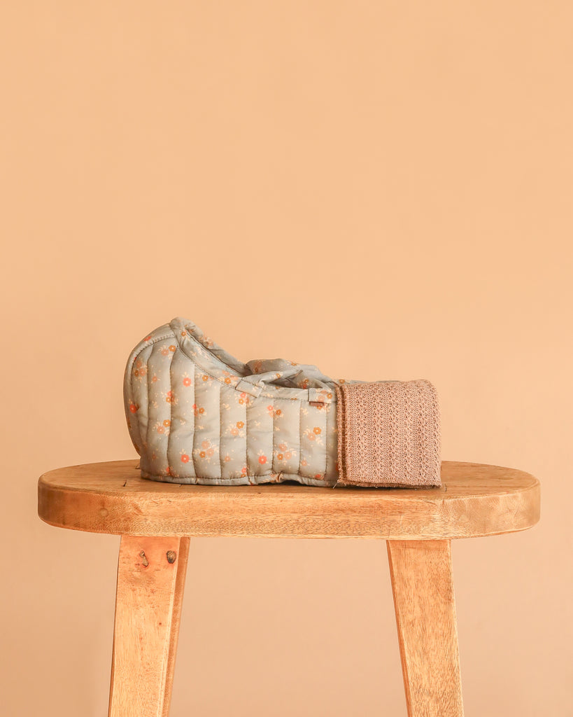 The Maileg Teddy Bear in Carry Cot, featuring a soft floral-patterned carrier with a knitted panel, is displayed on a wooden stool against a simple beige background.