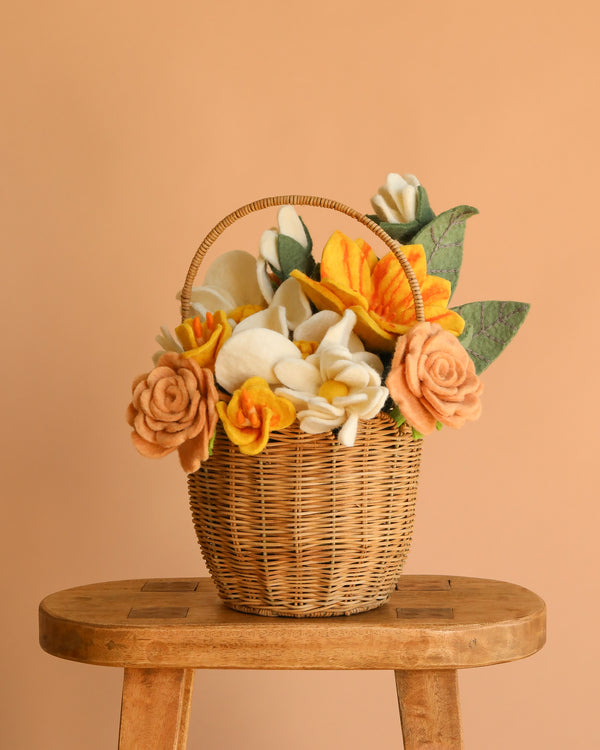 The Basket of Felt Flowers, filled with yellow, white, and peach felt blooms and green leaves, sits on a wooden stool against a peach-colored background.