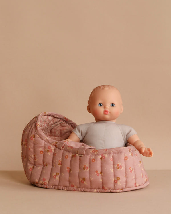 The Minikane Soft Body Doll (11") – Claire, with blue eyes and a grey outfit, sits upright in a pink floral-patterned Carry Cot bassinet against a beige background.