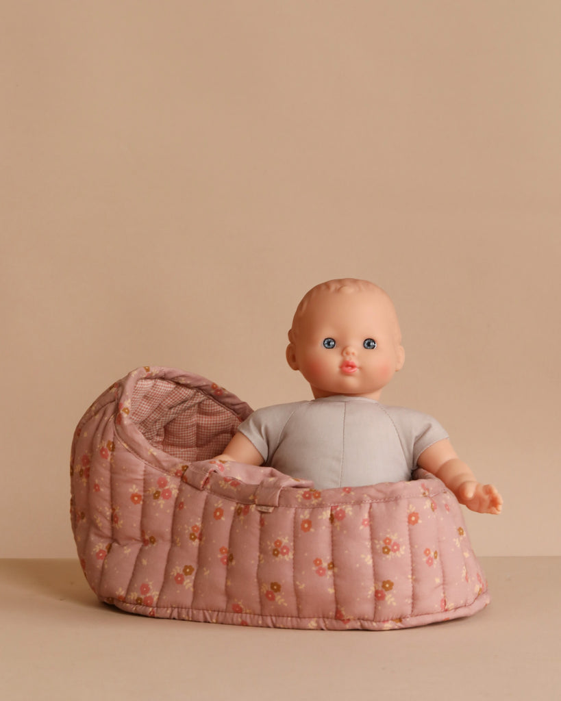 The Minikane Soft Body Doll (11") – Claire, with blue eyes and a grey outfit, sits upright in a pink floral-patterned Carry Cot bassinet against a beige background.
