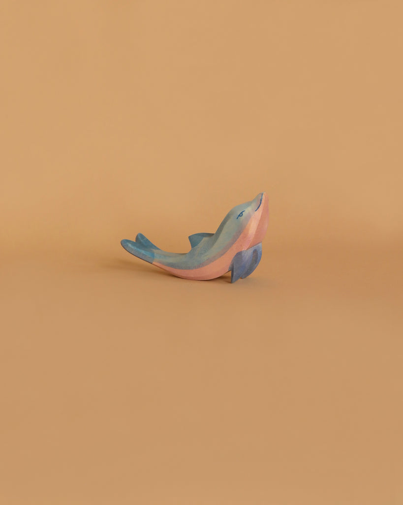 A small, smooth figurine resembling the Ostheimer Dolphin, Head High is displayed against a plain, warm beige background. Painted in shades of blue and pink, the dolphin is shown in a playful, leaping pose.