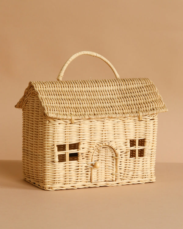 The Olli Ella Casa Bag is a hand-woven rattan basket shaped like a cozy house with a pitched roof, arched doorway, window details, and a top handle. Part of the Piki Baskets collection. Shown on a beige background.