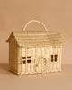 The Olli Ella Casa Bag is a hand-woven rattan basket shaped like a cozy house with a pitched roof, arched doorway, window details, and a top handle. Part of the Piki Baskets collection. Shown on a beige background.