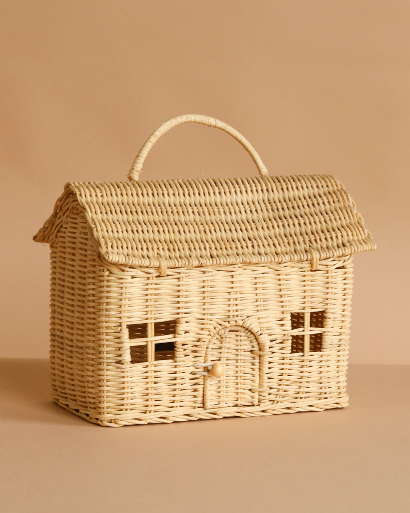 The Olli Ella Casa Bag is a hand-woven rattan basket shaped like a cozy house with a pitched roof, arched doorway, window details, and a top handle. Part of the Piki Baskets collection. Shown on a beige background.