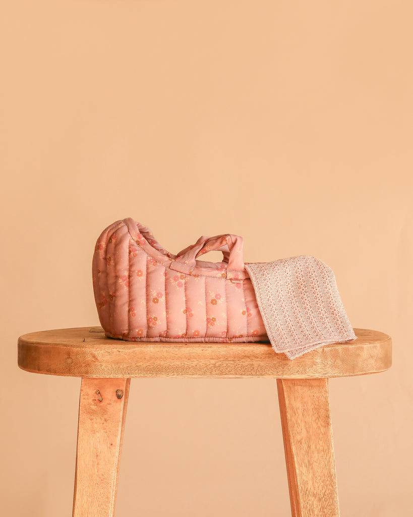 A Maileg Elephant in Carry Cot, featuring a small pink quilted bed with floral patterns and a knitted blanket, rests on a wooden stool against a plain beige background.